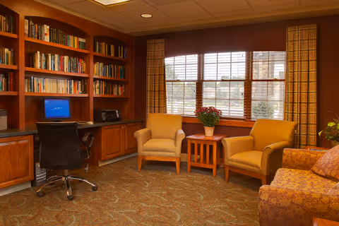 A cozy interior room with a built-in wooden bookshelf filled with books, a desk with a computer and printer, two beige armchairs around a small wooden table with a potted plant, and a patterned sofa. Large windows with blinds and curtains let in natural light.