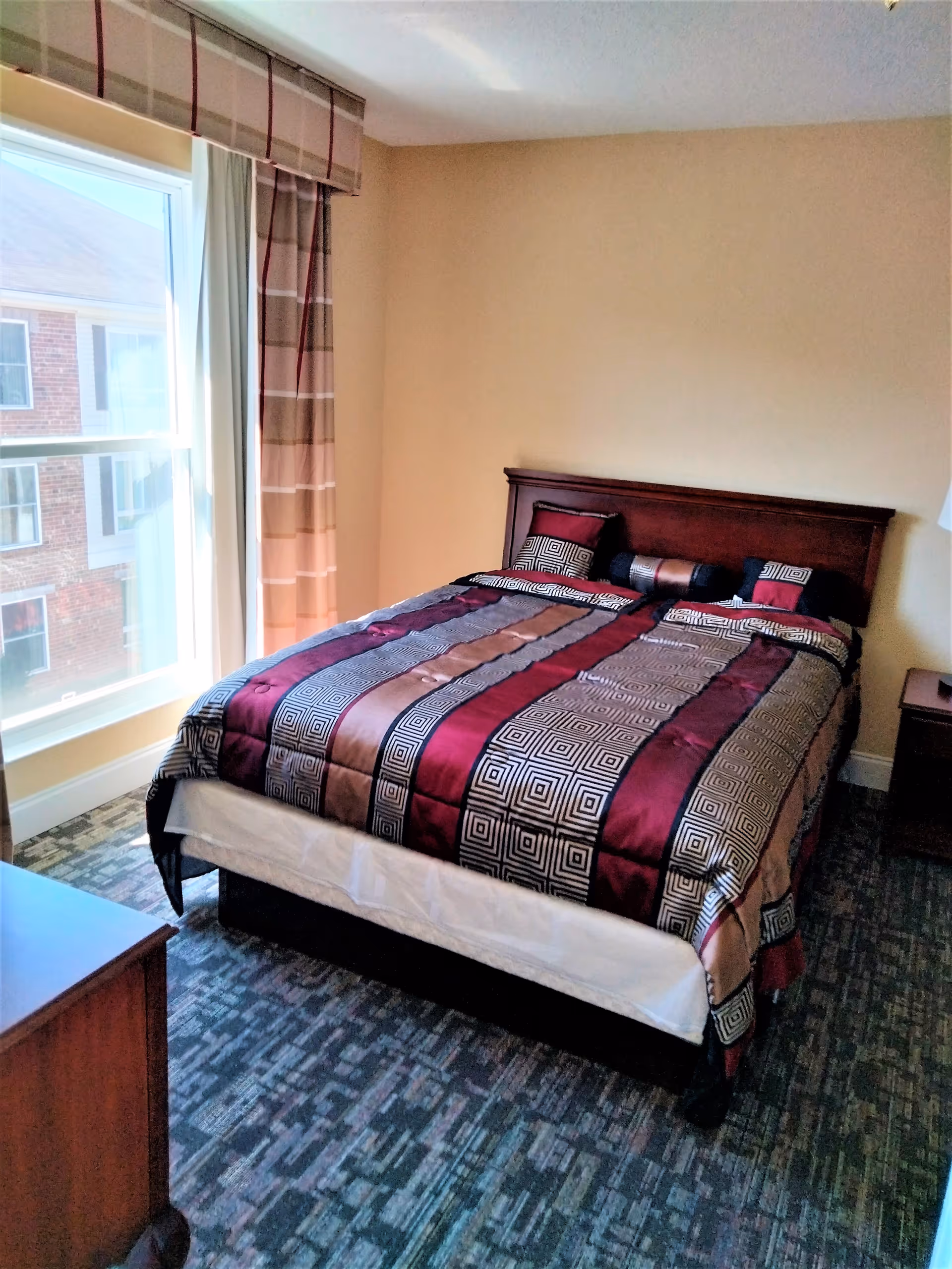 A bedroom with a large bed covered in a patterned bedspread with shades of burgundy, brown, and beige. The bed has matching pillows and a dark wooden headboard. There is a window with beige and brown plaid curtains letting in natural light. A wooden nightstand with a lamp is visible on the right side of the bed, and a wooden dresser is partially visible on the left side of the image. The floor is carpeted with a patterned design.