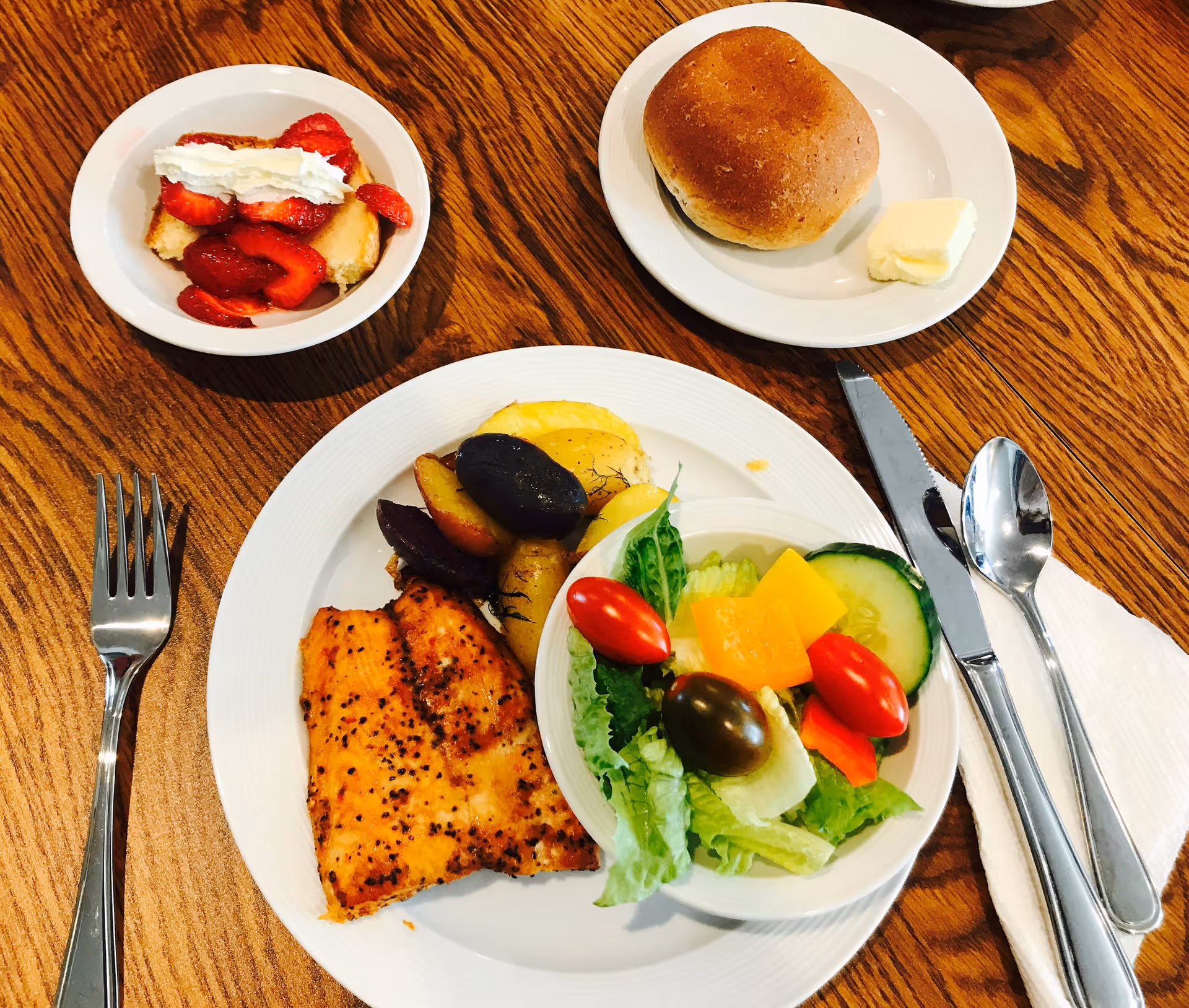 A meal on a wooden table consisting of a plate with grilled fish, roasted potatoes, and a small bowl of salad with lettuce, cherry tomatoes, cucumber, and cheese cubes. There is also a plate with a bread roll and a pat of butter, and a bowl of dessert topped with whipped cream and sliced strawberries. Silverware including a fork, knife, and spoon is placed on a napkin beside the plates.