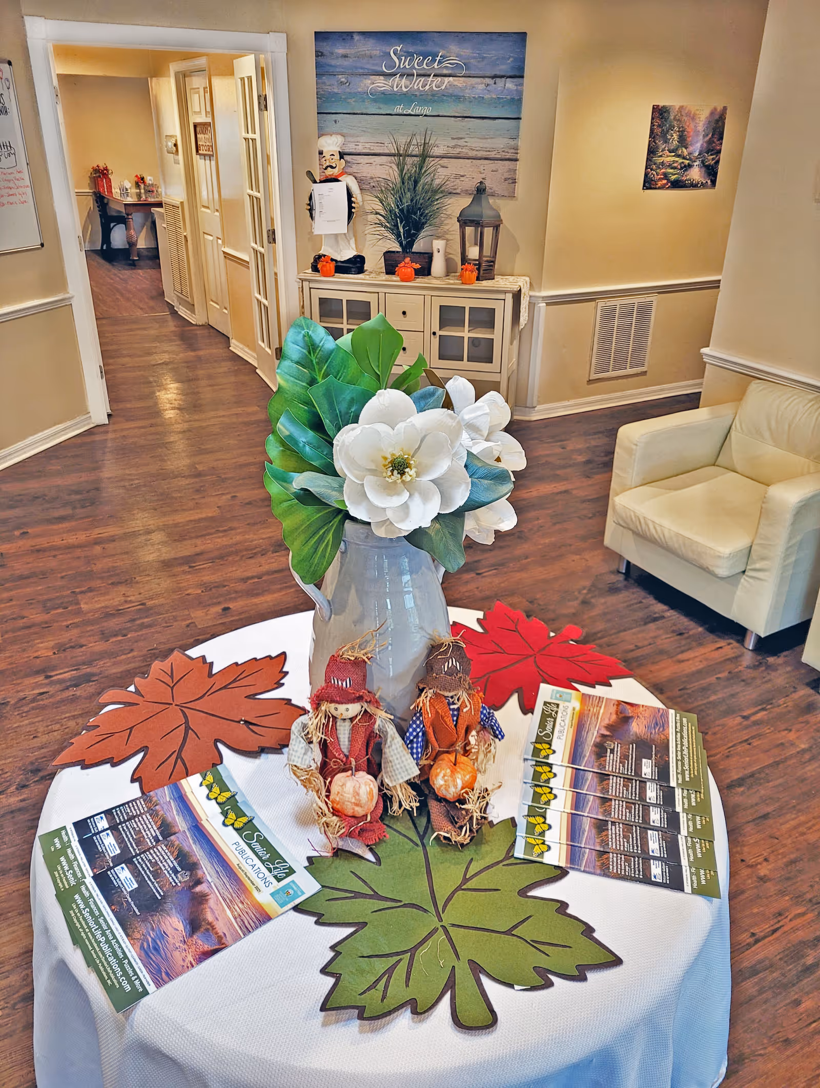Interior view of a senior living facility lobby area at Sweet Water at Largo, featuring a round table with a white tablecloth decorated with large autumn leaf placemats, a vase with white flowers and green leaves, two small scarecrow figurines holding pumpkins, and brochures. In the background, there is a cream-colored armchair, a cabinet with decorative items including a chef figurine, small pumpkins, a plant, and a lantern. A sign on the wall reads 'Sweet Water at Largo'. The floor is wooden and the walls are painted beige.