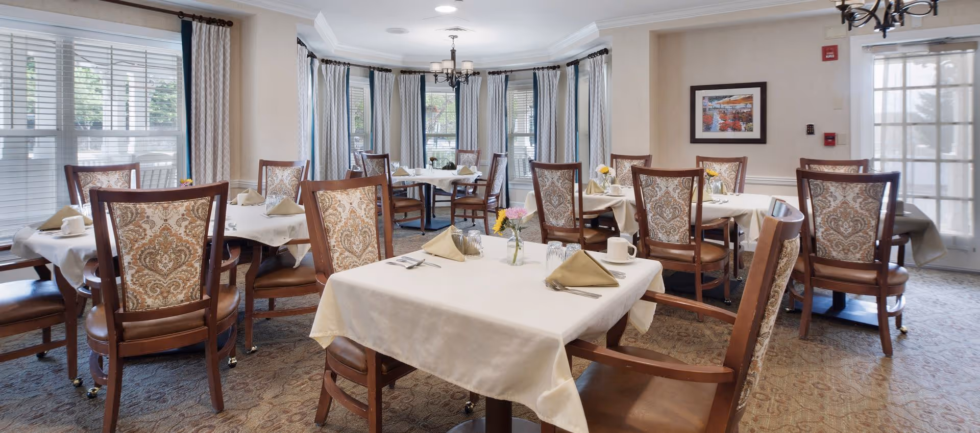 A bright dining room with several tables covered in white tablecloths, each set with beige napkins, cups, glasses, and silverware. The chairs have wooden frames with patterned upholstery. Large windows with white blinds and curtains allow natural light to fill the room. A chandelier hangs from the ceiling, and a framed picture is on the wall near a glass door.