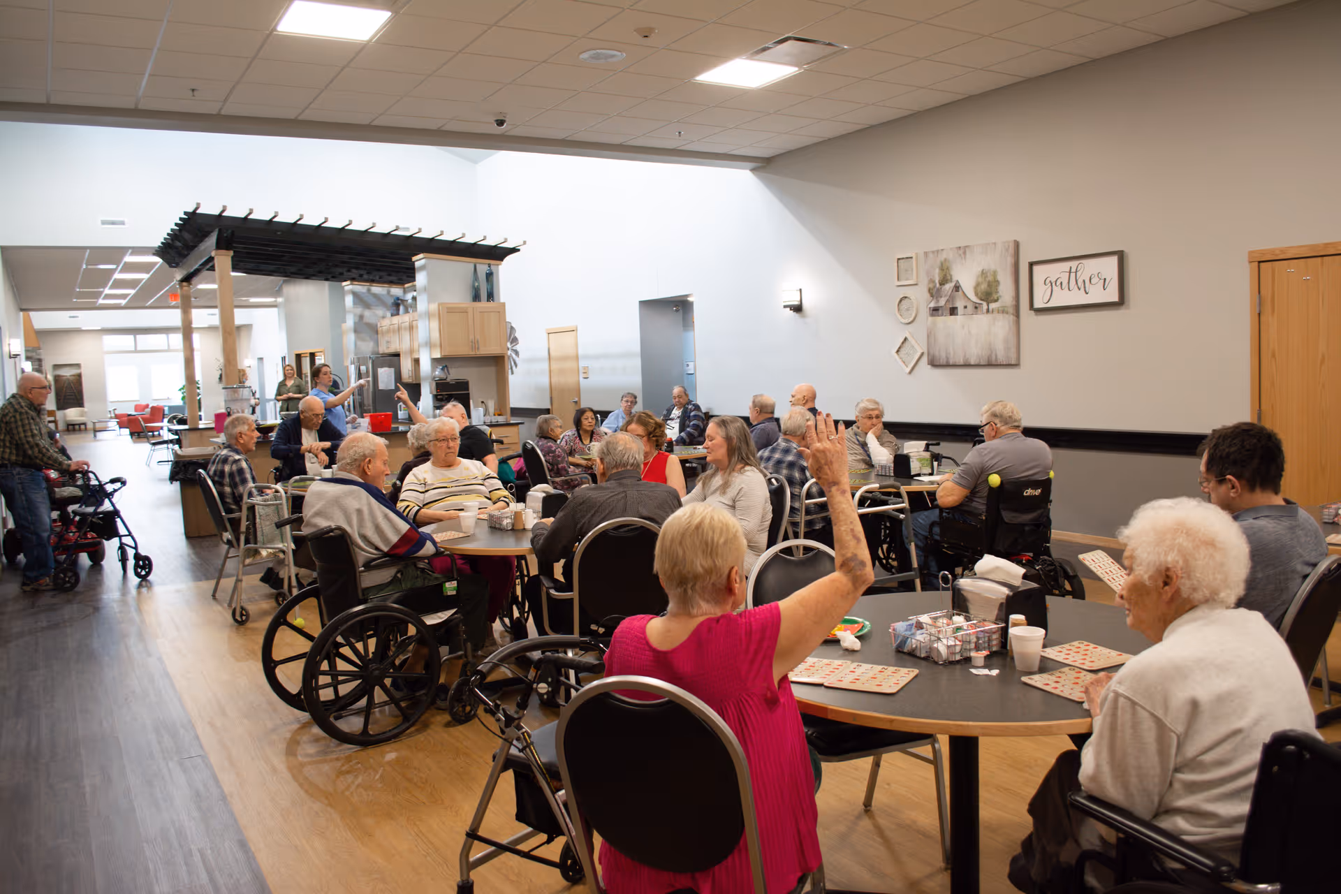 A group of elderly people sitting around tables in a large, well-lit room playing bingo. Some individuals are in wheelchairs, and one woman in a pink top has her hand raised. The room has light-colored walls with framed artwork and a sign that says 'gather'. There is a kitchen area in the background with a pergola-style structure above it.