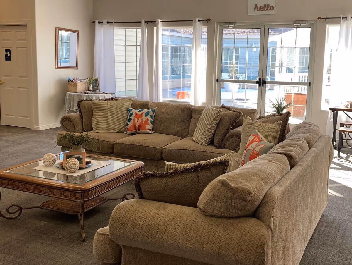 A cozy living room area with two beige sofas arranged around a glass-top wooden coffee table. The sofas have various decorative pillows, including one with a fox design. Behind the sofas are large windows and glass doors letting in natural light, with white curtains. A small table with a basket and some items is against the wall near a door marked as a restroom. A small 'hello' sign is mounted above the glass doors.