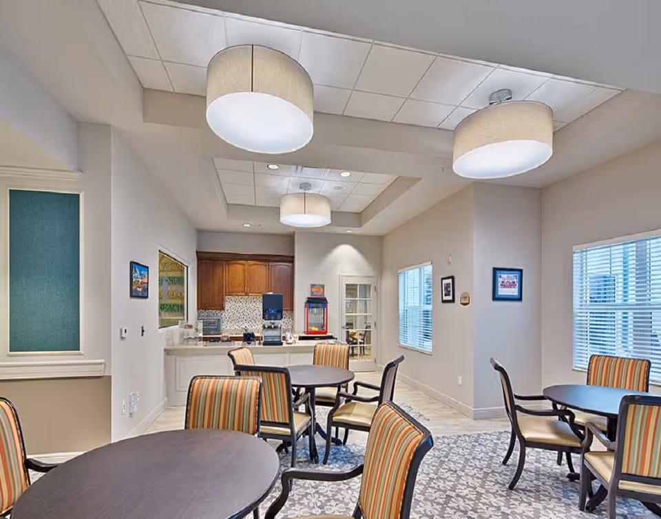 Interior view of a dining area in Lexington Place featuring round tables with striped cushioned chairs, large ceiling lights, windows with blinds, and a counter area with a popcorn machine and coffee dispenser in the background.