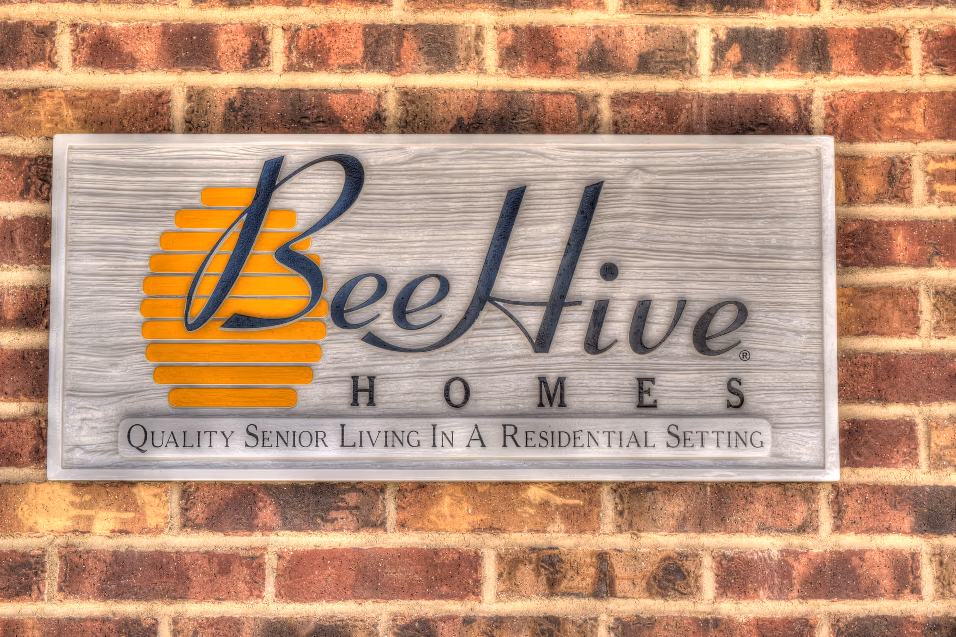 A wooden sign mounted on a brick wall displaying the logo and name 'BeeHive Homes' with the tagline 'Quality Senior Living In A Residential Setting'. The logo features a stylized bee hive in orange.