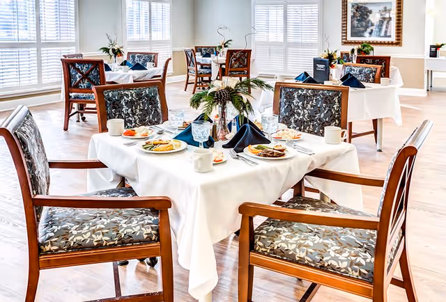 A bright dining room with wooden chairs featuring floral patterned cushions around tables covered with white tablecloths. Each table is set with plates of food, glasses, mugs, and napkins folded in a decorative manner. Large windows with white blinds allow natural light to fill the room, and a framed picture hangs on the wall.