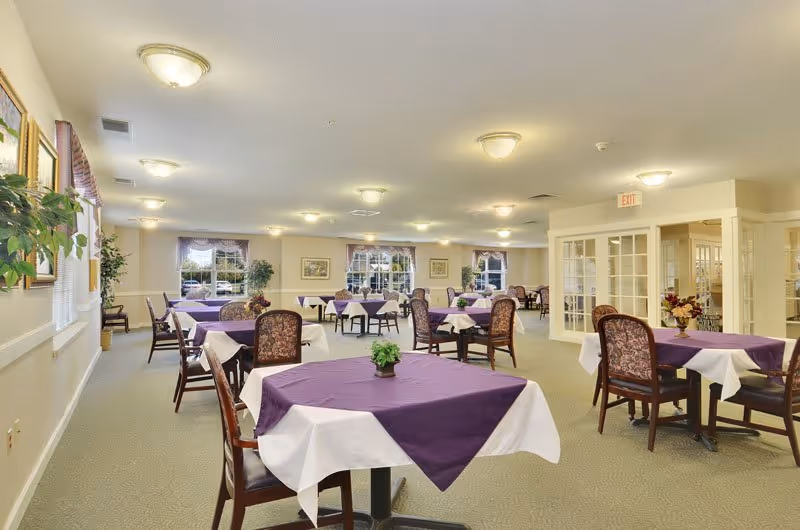 Spacious senior dining room with multiple tables covered in purple tablecloths and chairs arranged under ceiling lights.