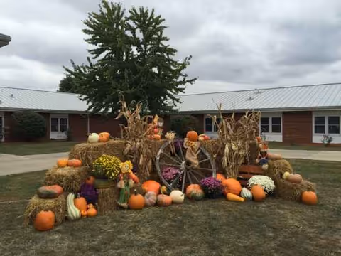Outdoor autumn display with pumpkins, hay bales, corn stalks, and a wagon wheel in front.
