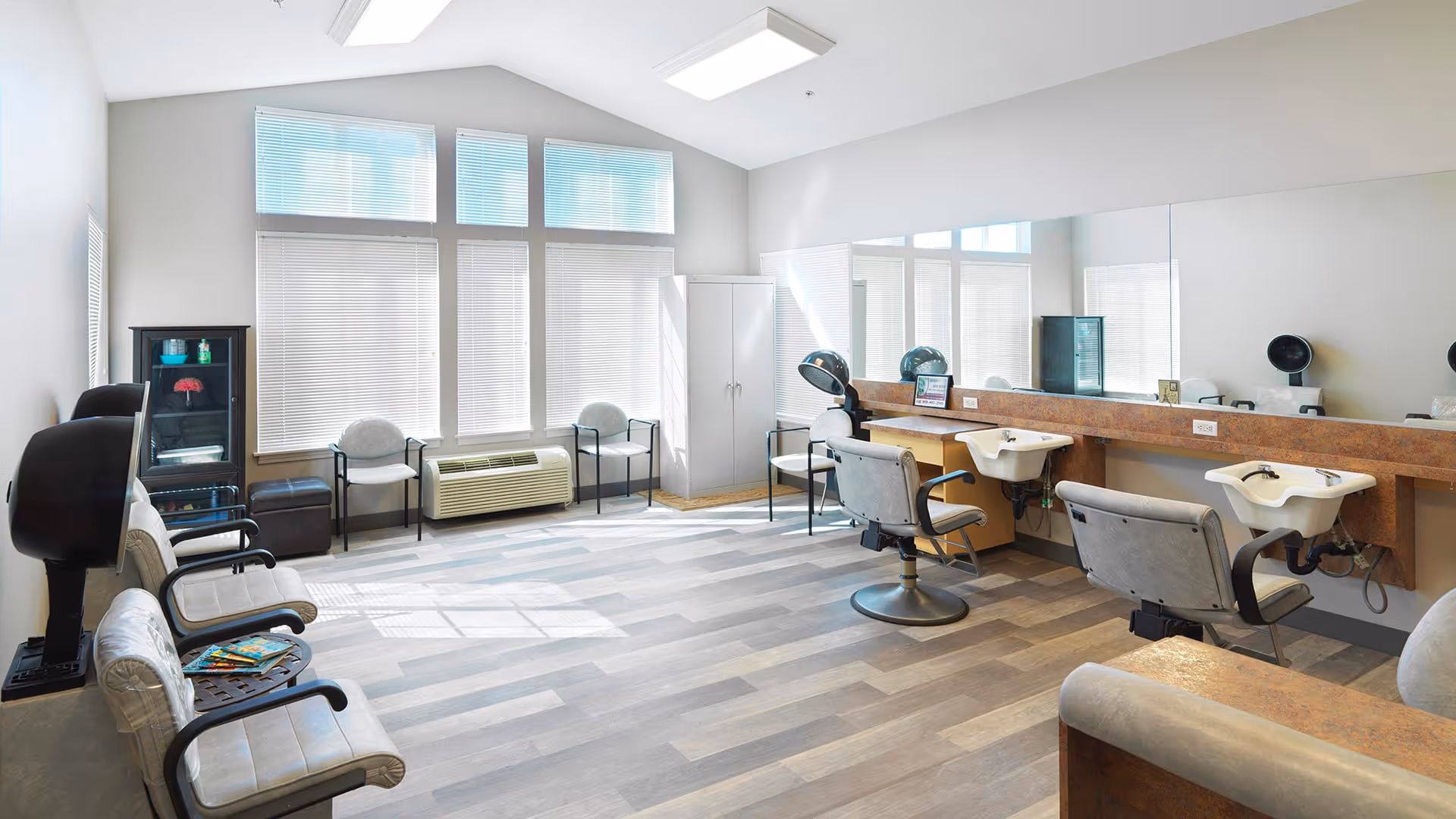 A bright and spacious hair salon area with large windows covered by blinds allowing natural light to fill the room. The space features salon chairs with attached sinks for washing hair, a large mirror along one wall, waiting chairs with armrests, a small cabinet with hair products, and a hair dryer station. The floor has a wood-like pattern, and the walls are painted light gray.