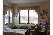 A cozy room with a bed in the foreground, a patterned sofa against the wall under two large windows with curtains, and various decorations on the walls including pictures and a clock.