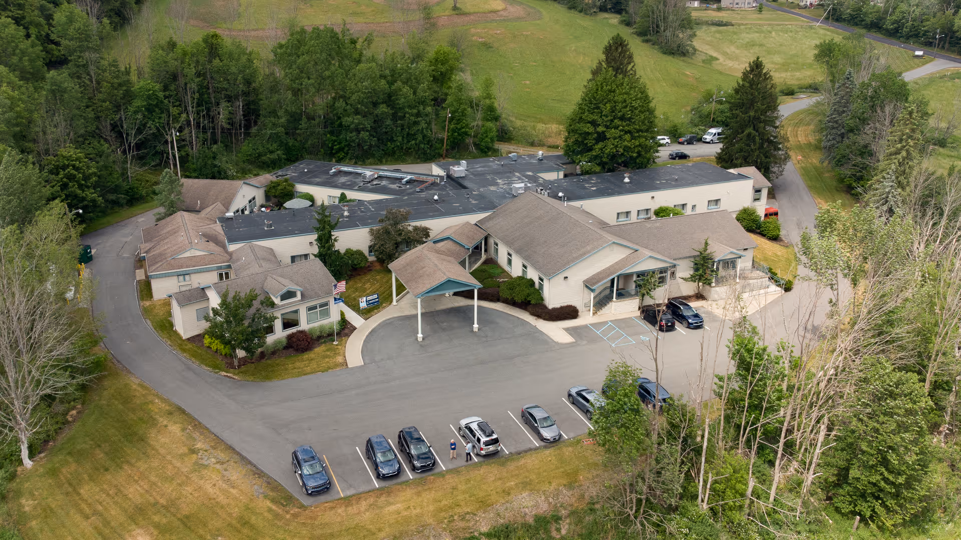 Aerial view of Meadow View Rehabilitation and Healthcare Center, showing a single-story building with a covered entrance, surrounded by trees and greenery. Several cars are parked in the parking lot in front of the building.