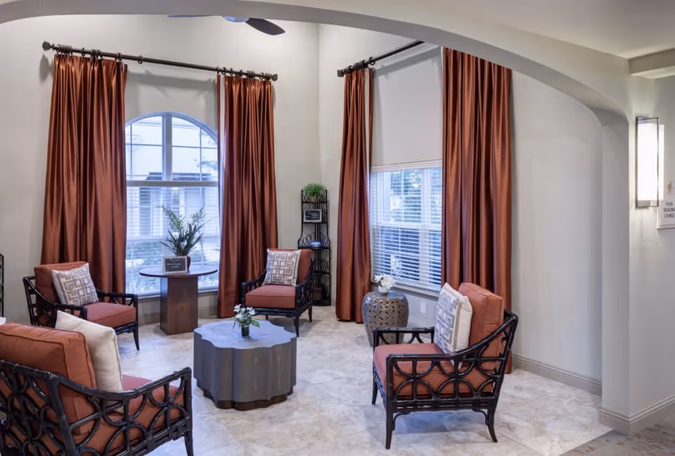 A cozy seating area in a senior living facility with four cushioned chairs arranged around a decorative coffee table. The room features large windows with rust-colored curtains, a small round table with a plant, and a light-colored tiled floor. The walls are painted light gray, and there is a wall-mounted light fixture on the right side.