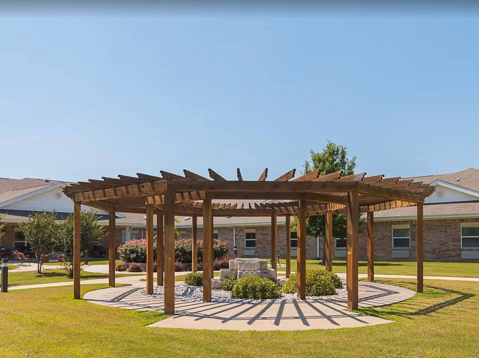 Outdoor circular wooden pergola structure with a stone fire pit in the center, surrounded by green grass and bushes. In the background, there are single-story brick buildings with windows and a clear blue sky.