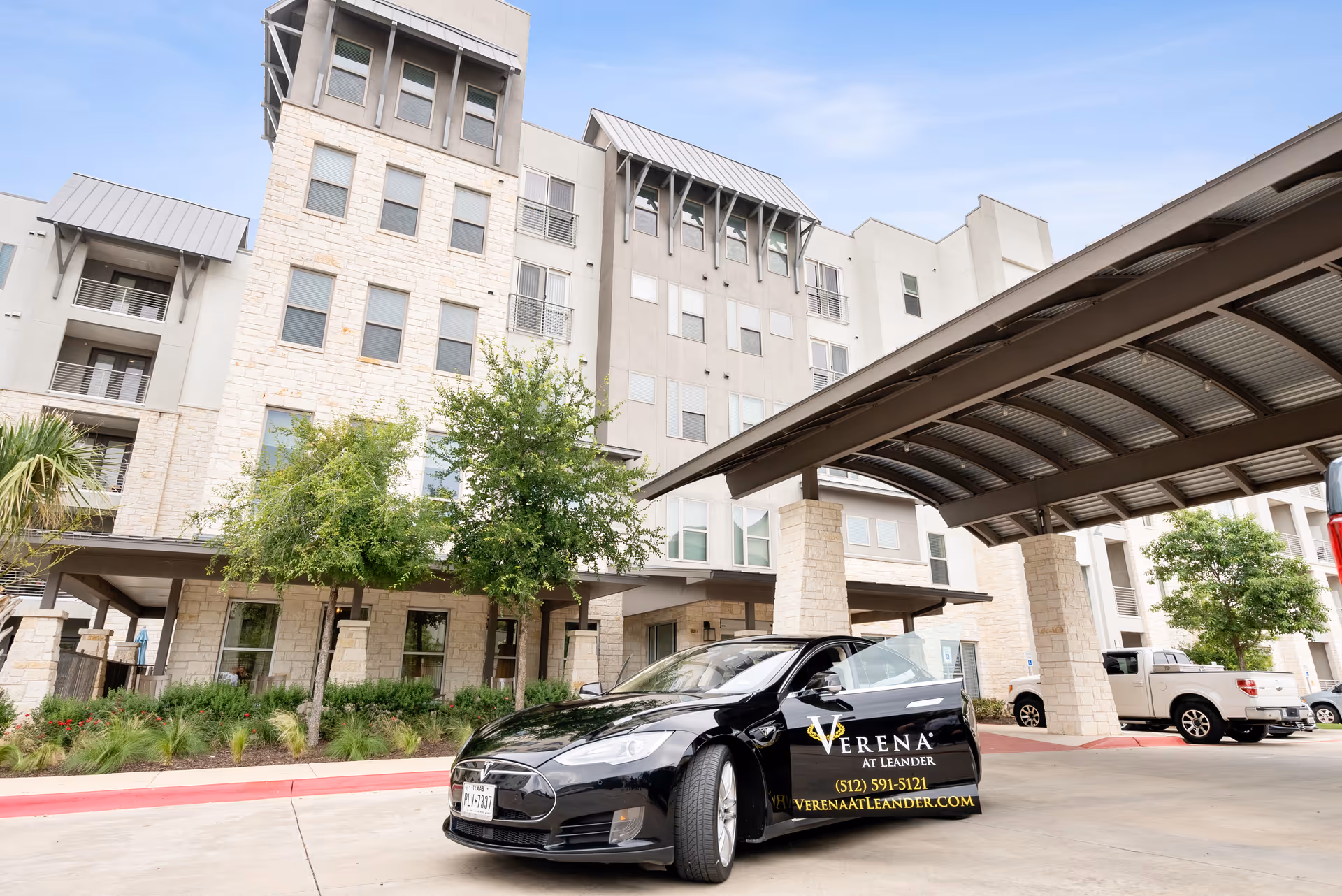 Exterior view of a multi-story senior living facility with stone and beige walls, trees, and a covered driveway. A black car with the Verena at Leander logo and contact information is parked in front.