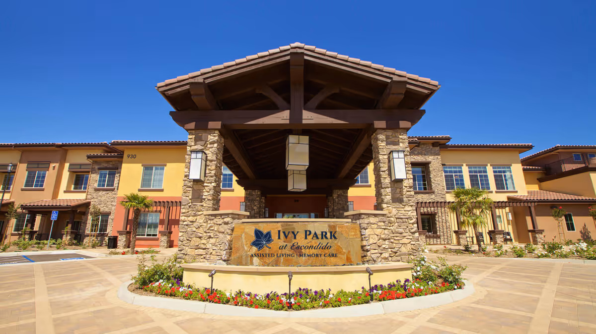 Front exterior view of Ivy Park at Escondido, an assisted living and memory care facility, featuring a large covered entrance with stone pillars, a sign with the facility name, and a landscaped area with flowers under a clear blue sky.
