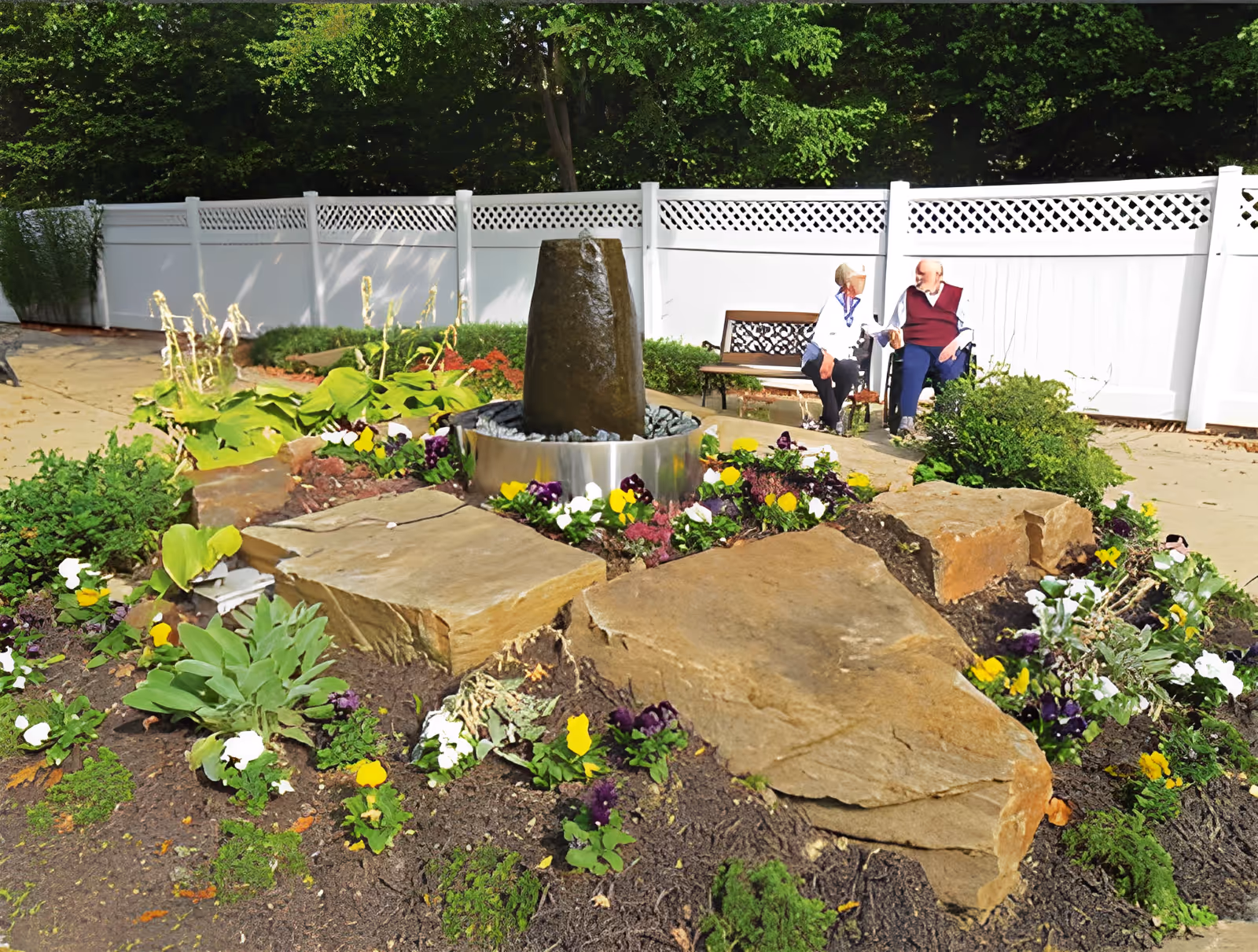A garden area with a stone water fountain surrounded by colorful flowers and large rocks. In the background, two elderly people are sitting on a bench and a wheelchair near a white fence, engaged in conversation.