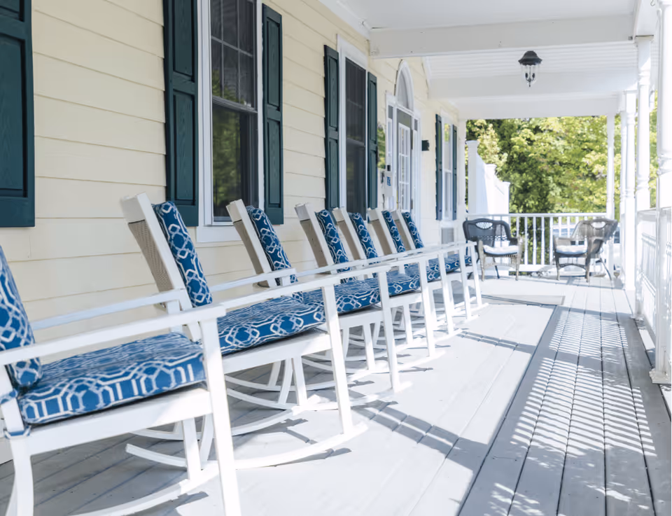 Sunny front porch with a row of white rocking chairs with blue patterned cushions along a yellow-sided building.