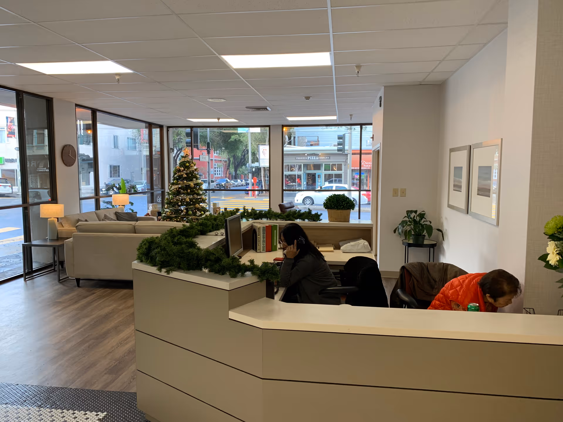 Interior view of a reception area in a senior living facility with a front desk decorated with green garland. Two people are seated behind the desk, one talking on the phone and the other looking down. In the background, there is a Christmas tree, large windows showing a street view, and a seating area with sofas and lamps.