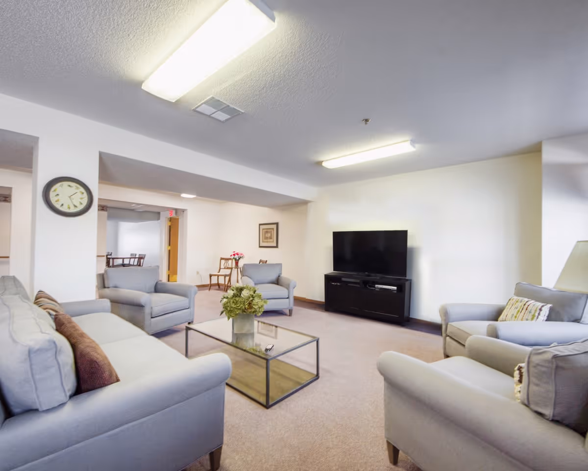 A cozy living room with light gray sofas and armchairs arranged around a glass coffee table with a flower vase. A flat-screen TV is placed on a black TV stand against the wall. The room has beige carpet, white walls, a wall clock, and overhead fluorescent lighting.