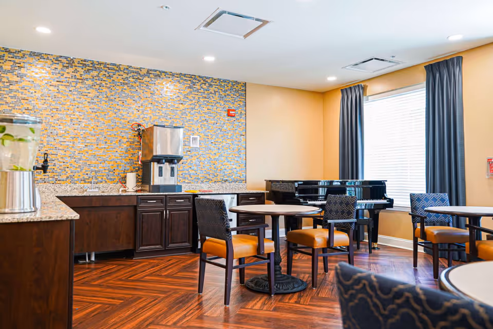 A bright room with wooden flooring and yellow walls featuring a small kitchenette area with a water dispenser and sink. There are round tables with chairs that have orange seats and patterned backs. A black grand piano is positioned near a window with blue curtains.