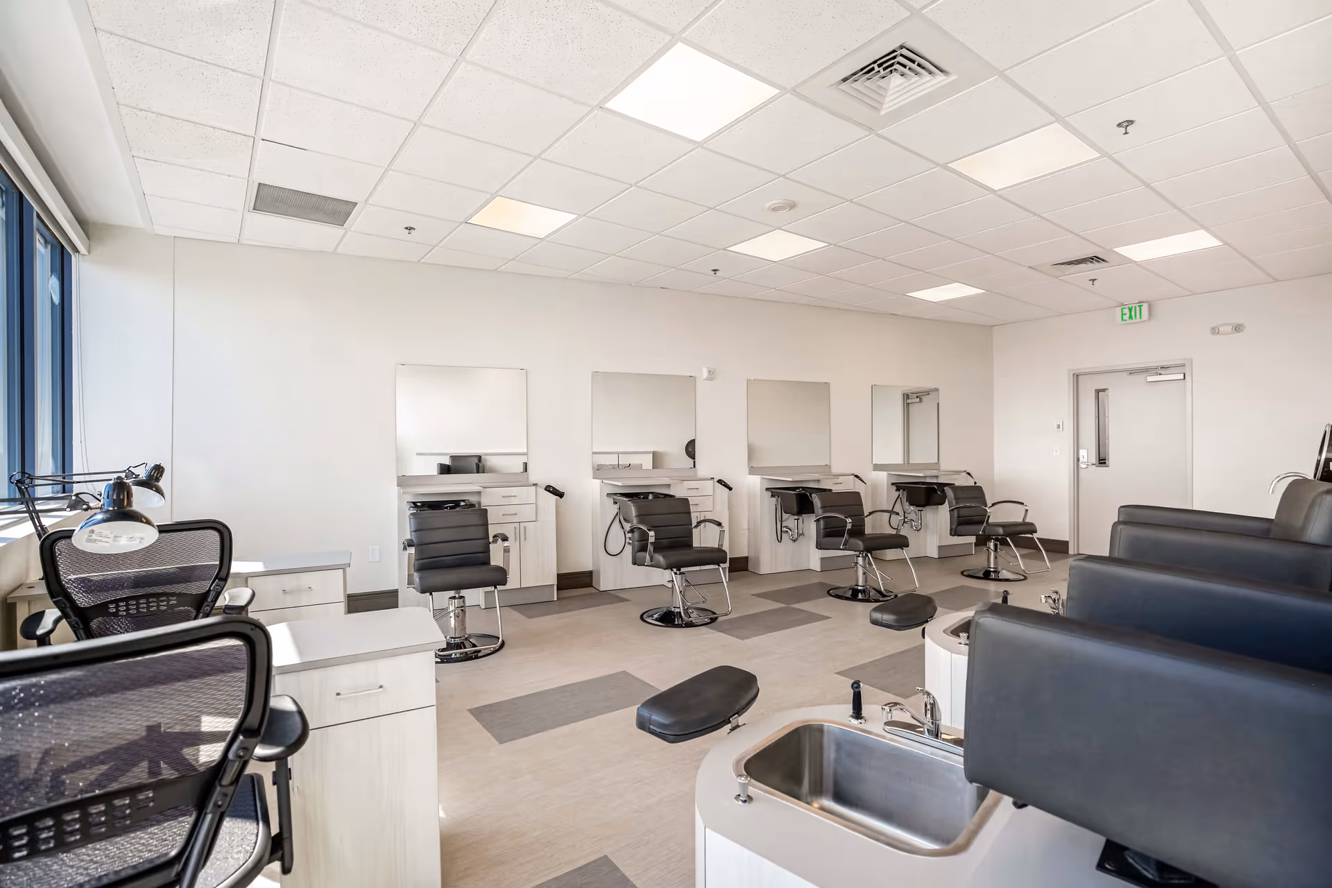 Interior view of a modern salon area in Copeland Tower Living with multiple black salon chairs in front of mirrors along the wall, pedicure stations with black chairs and foot baths, and office chairs near desks by large windows.