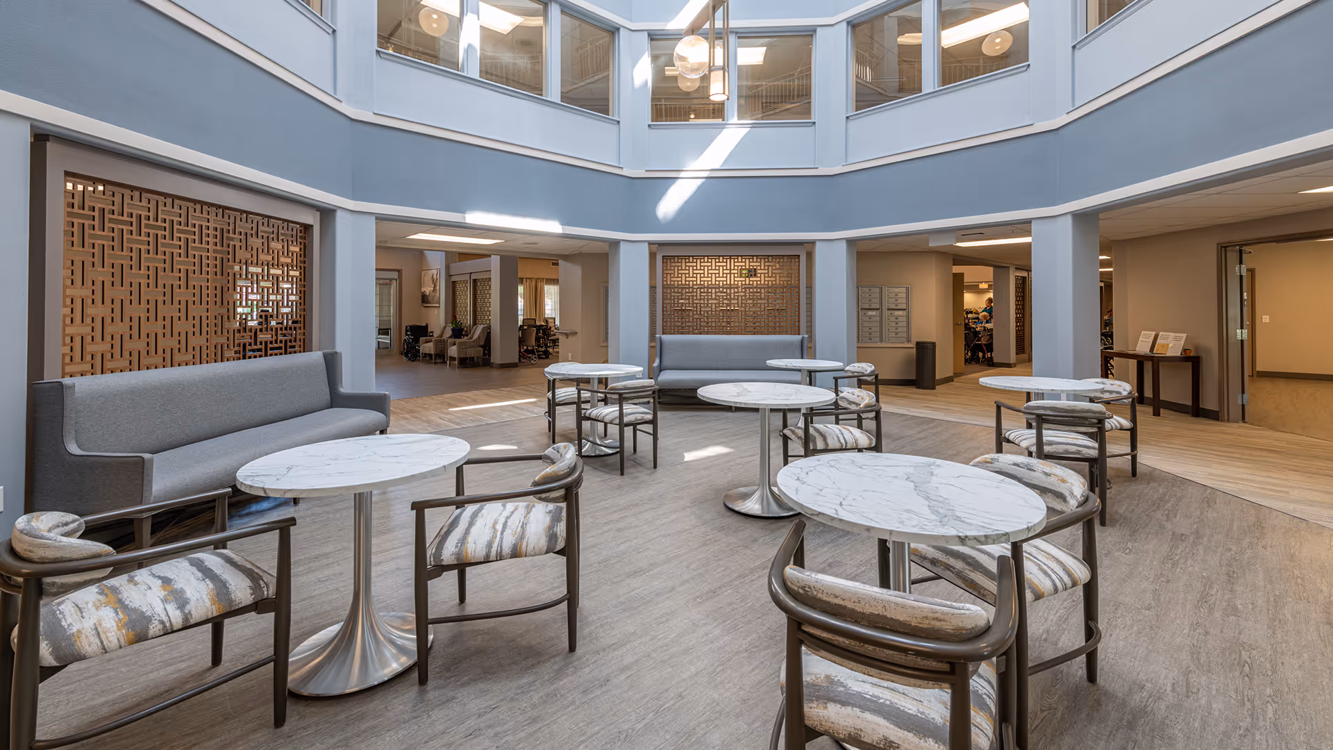 A bright, modern indoor common area with round marble-top tables and cushioned chairs arranged around the space. There are two gray upholstered benches against the walls with decorative wooden lattice panels behind them. The area is well-lit with natural light coming from large windows above, and the flooring is a light wood tone.