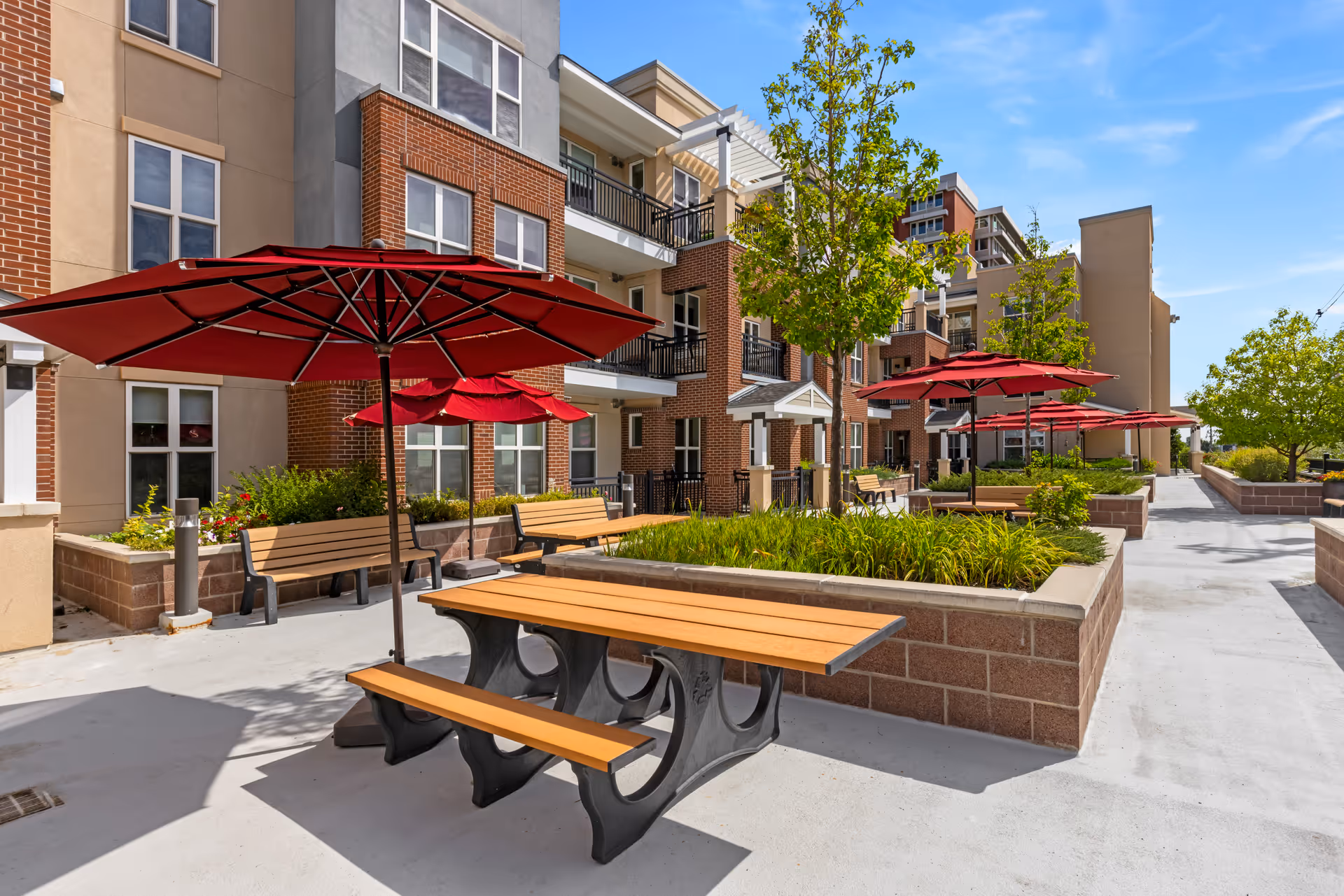 Outdoor courtyard with picnic tables, red umbrellas, benches and landscaping in front of a multi-story senior living building.