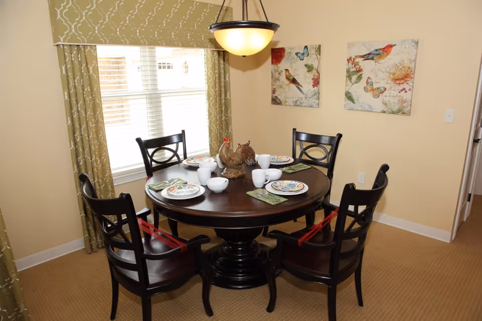 A dining area with a round dark wooden table set for four people. Each place setting includes a plate, a bowl, a cup, and a green napkin. Four dark wooden chairs with armrests surround the table, and two of the chairs have red straps attached. The room has beige walls, a window with green patterned curtains, and two framed pictures of birds and butterflies on the wall. A hanging light fixture is above the table.