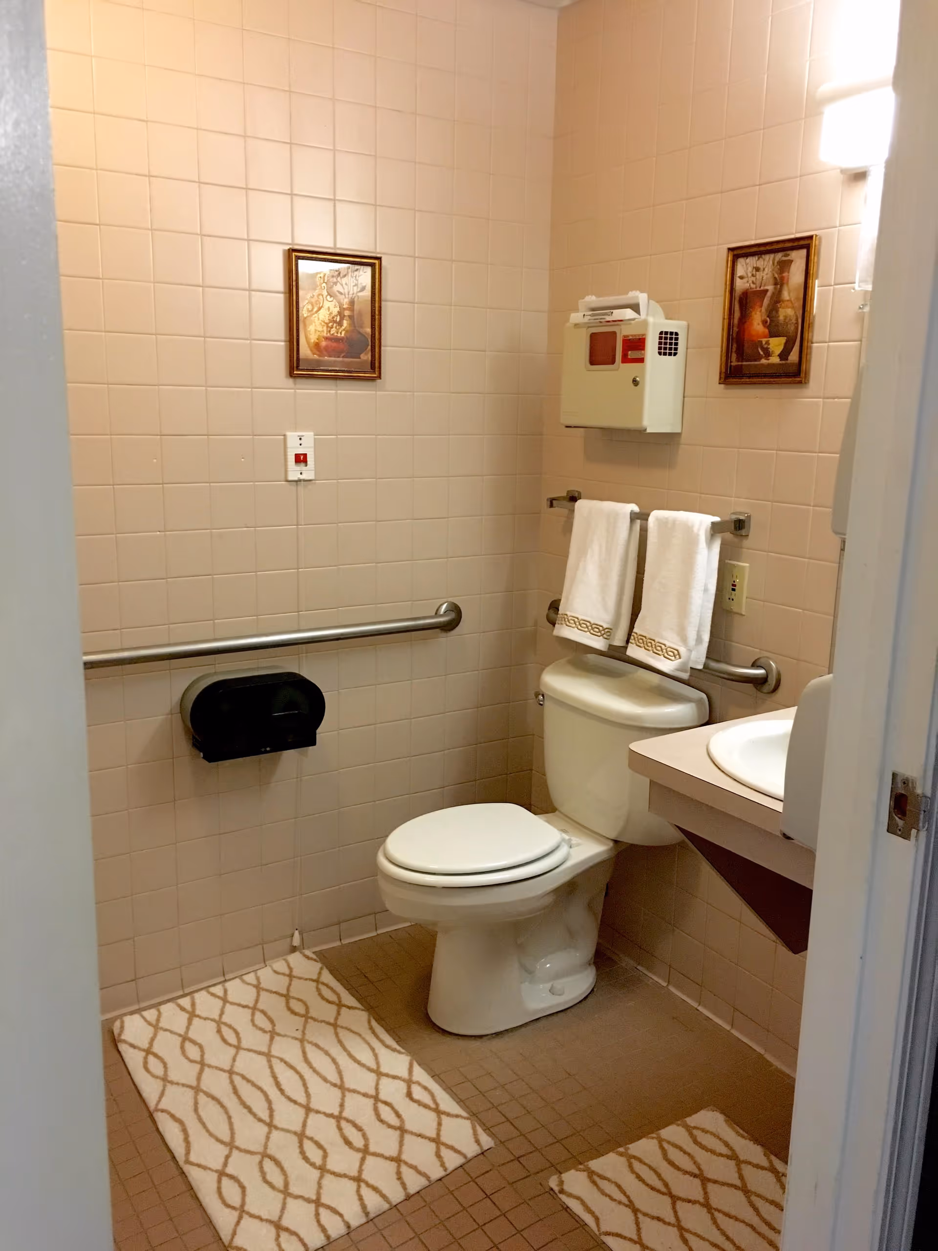 A small bathroom with beige tiled walls and floor. It features a white toilet with two white towels hanging on a rail above the tank. There are two framed pictures on the walls, a white sink with a countertop, a wall-mounted soap dispenser, a grab bar next to the toilet, and a beige patterned bath mat on the floor.
