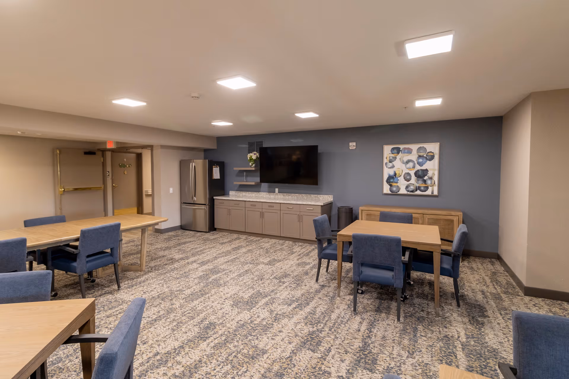 A spacious common area with multiple wooden tables and blue upholstered chairs arranged around them. The room has a patterned carpet floor, beige walls, and a dark blue accent wall. On the accent wall, there is a large flat-screen TV mounted above a long cabinet with a granite countertop. A stainless steel refrigerator is positioned in the corner, and a piece of abstract art hangs on the wall next to the TV. The ceiling has recessed lighting, and there is an open doorway leading to another area.