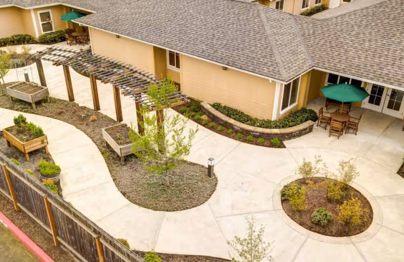 Aerial view of a courtyard with curved concrete walkways, raised planters, a wooden pergola, and outdoor seating next to a beige building.