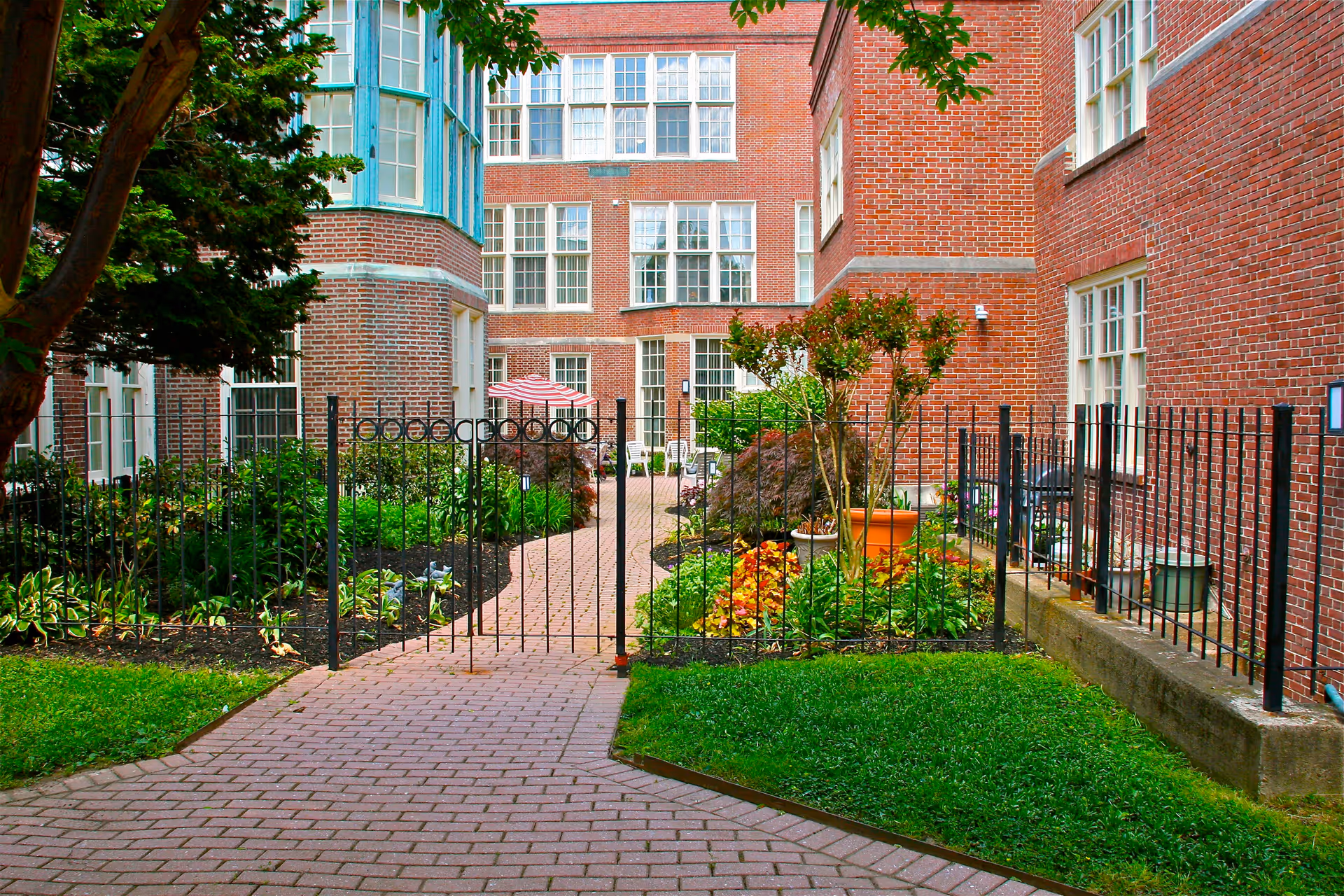 A gated garden courtyard area surrounded by red brick buildings with multiple windows. The courtyard features a paved walkway, green grass, various plants, and a patio umbrella with chairs in the background.