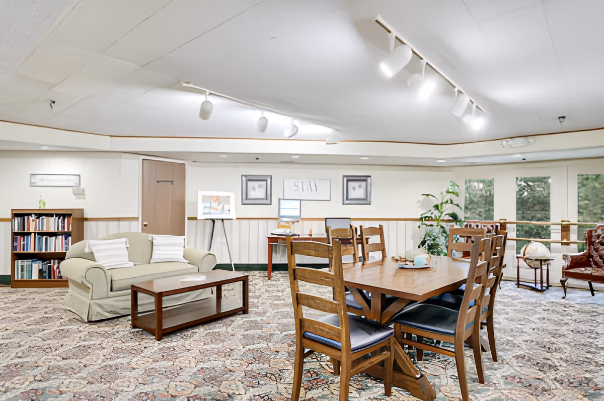 Spacious senior living common room with a sofa, coffee table, bookshelf, and a wooden dining table with chairs.