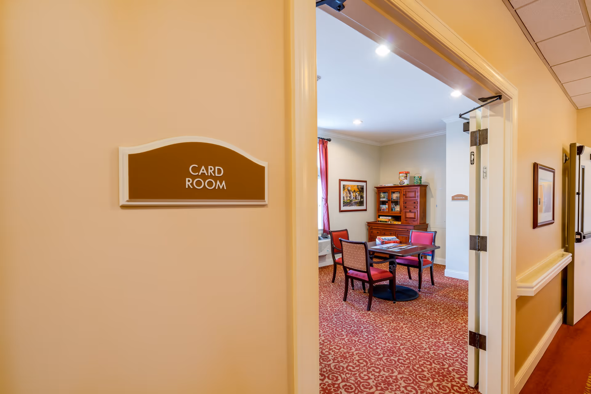 View through an open door into a card room with a table and four chairs. The room has patterned red carpet, a wooden cabinet with games and decorations, and a window with red curtains. A sign on the wall outside the door reads 'CARD ROOM'.