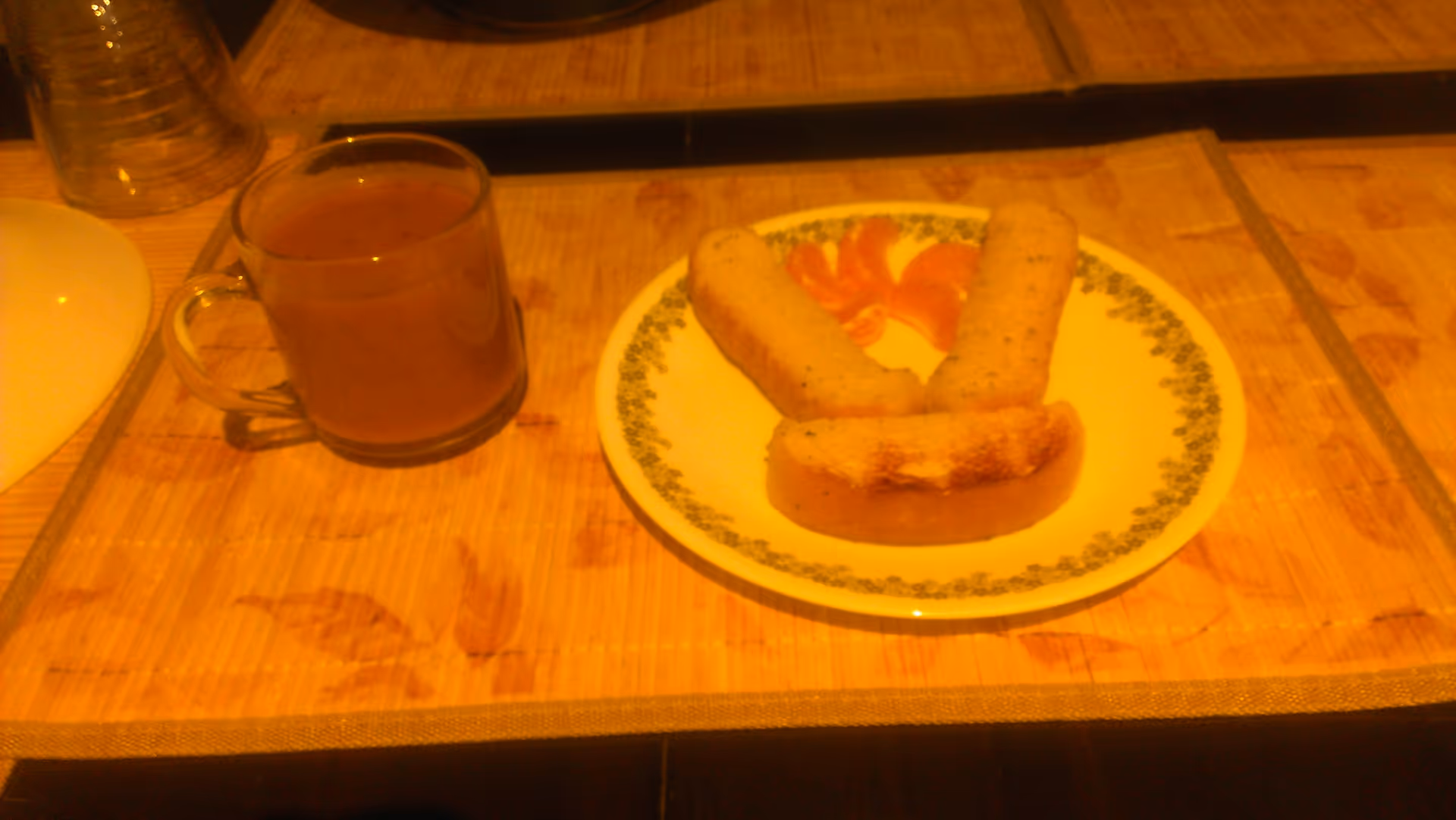 A clear glass cup filled with a brown liquid, placed on a bamboo placemat next to a white plate with a green floral border. The plate contains three pieces of toasted bread arranged in a smiley face pattern with orange slices as eyes.