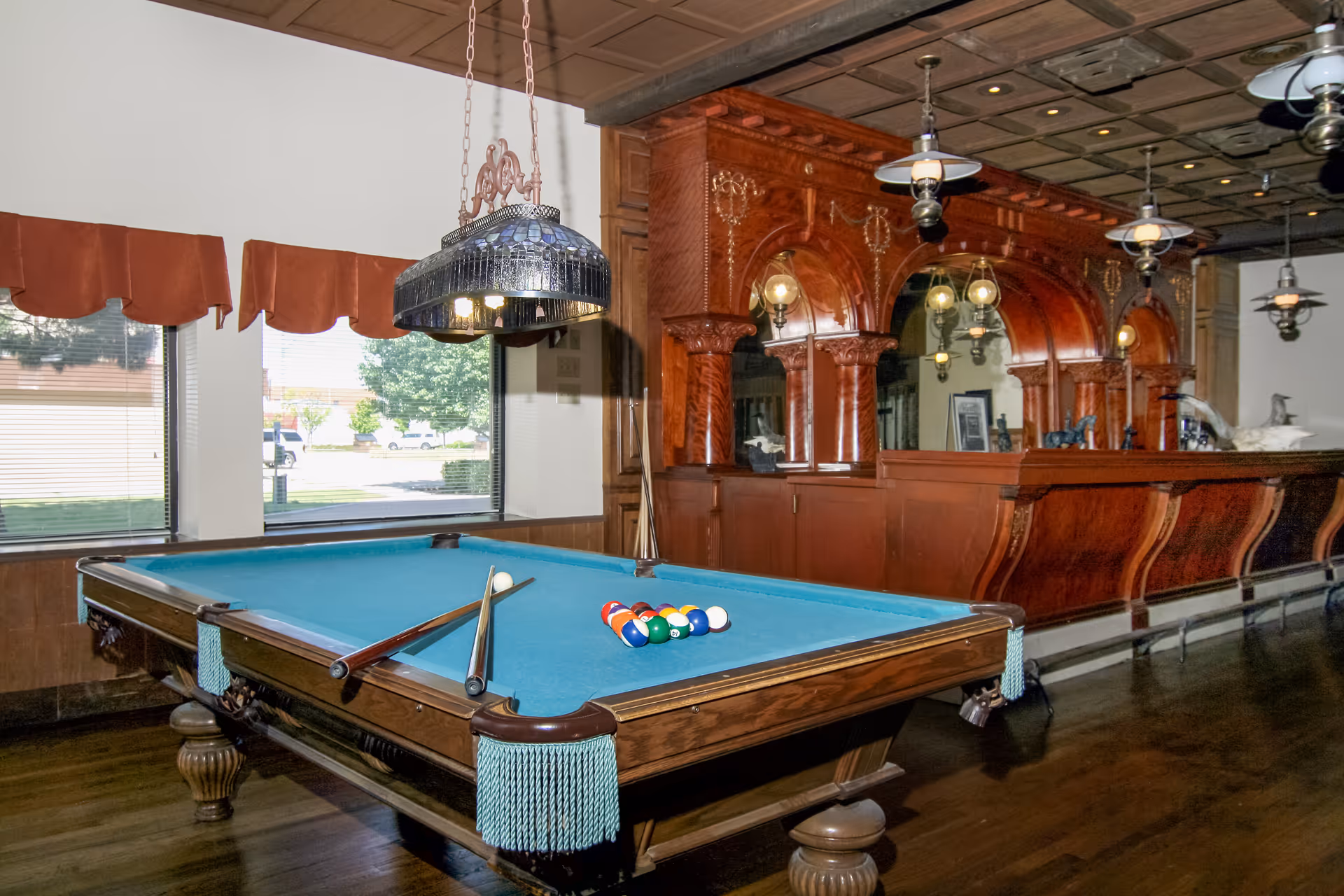 A billiards room with a pool table set up for a game, featuring a blue felt surface, pool cues resting on the table, and a rack of colorful billiard balls. The room has large windows with red valances, a wooden floor, and an ornate wooden bar with mirrors and hanging lights in the background.