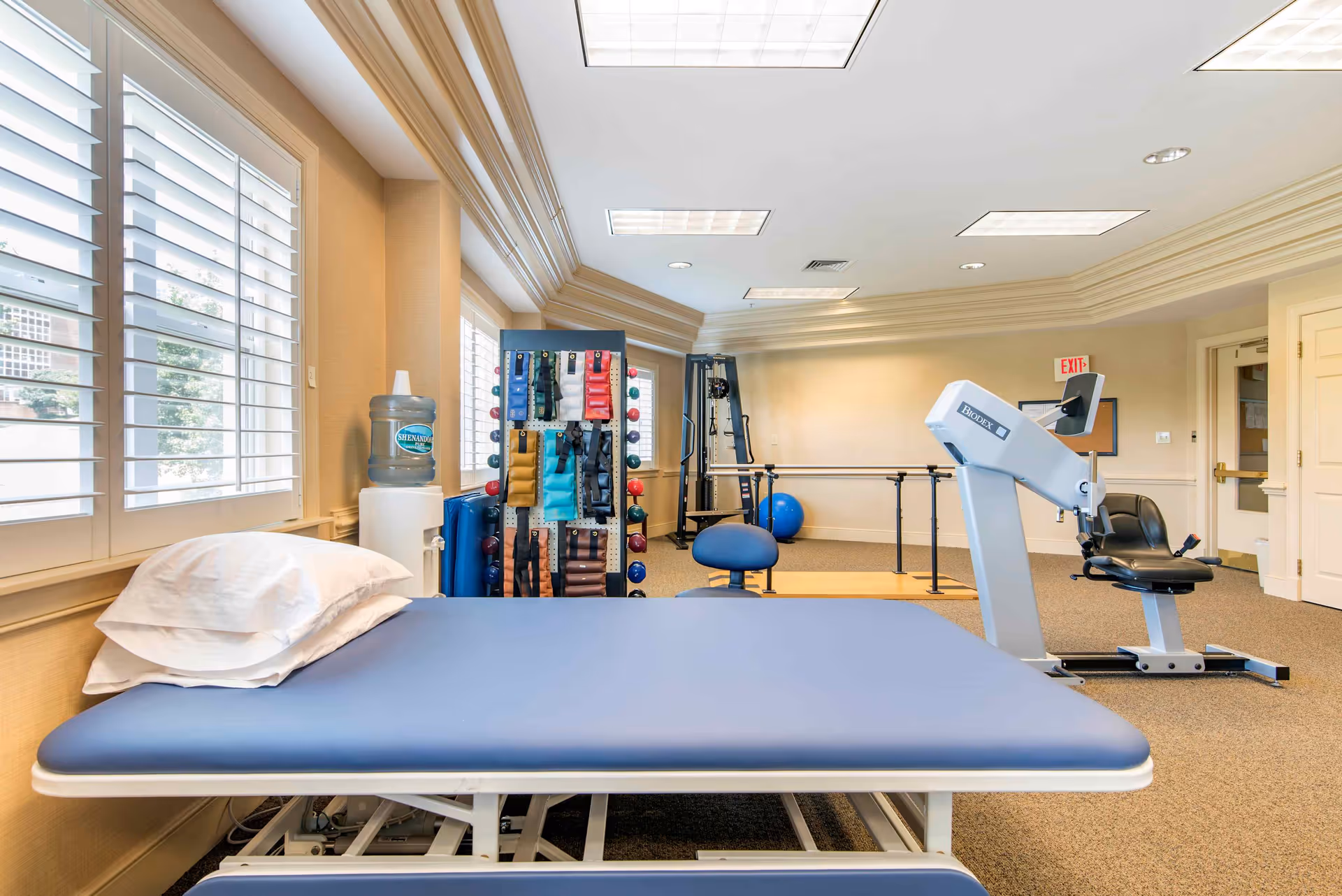 A bright physical therapy or rehabilitation room with a blue padded treatment table and pillow in the foreground. The room has large windows with white shutters letting in natural light. In the background, there is exercise equipment including a recumbent bike, parallel bars, a rack with various resistance bands and weights, and an exercise ball. The walls are light-colored, and the ceiling has recessed lighting panels.