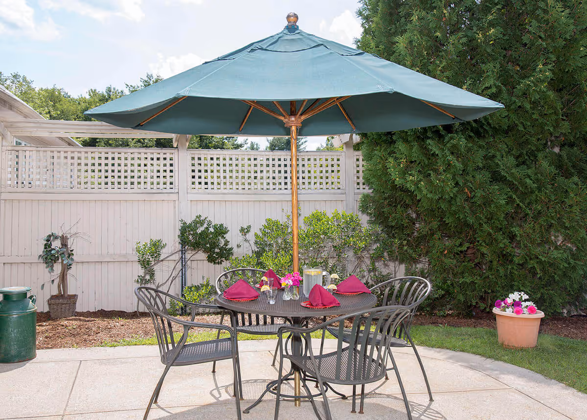 Patio dining set with a large green umbrella over a metal table and chairs set with napkins and flowers in a fenced garden area.