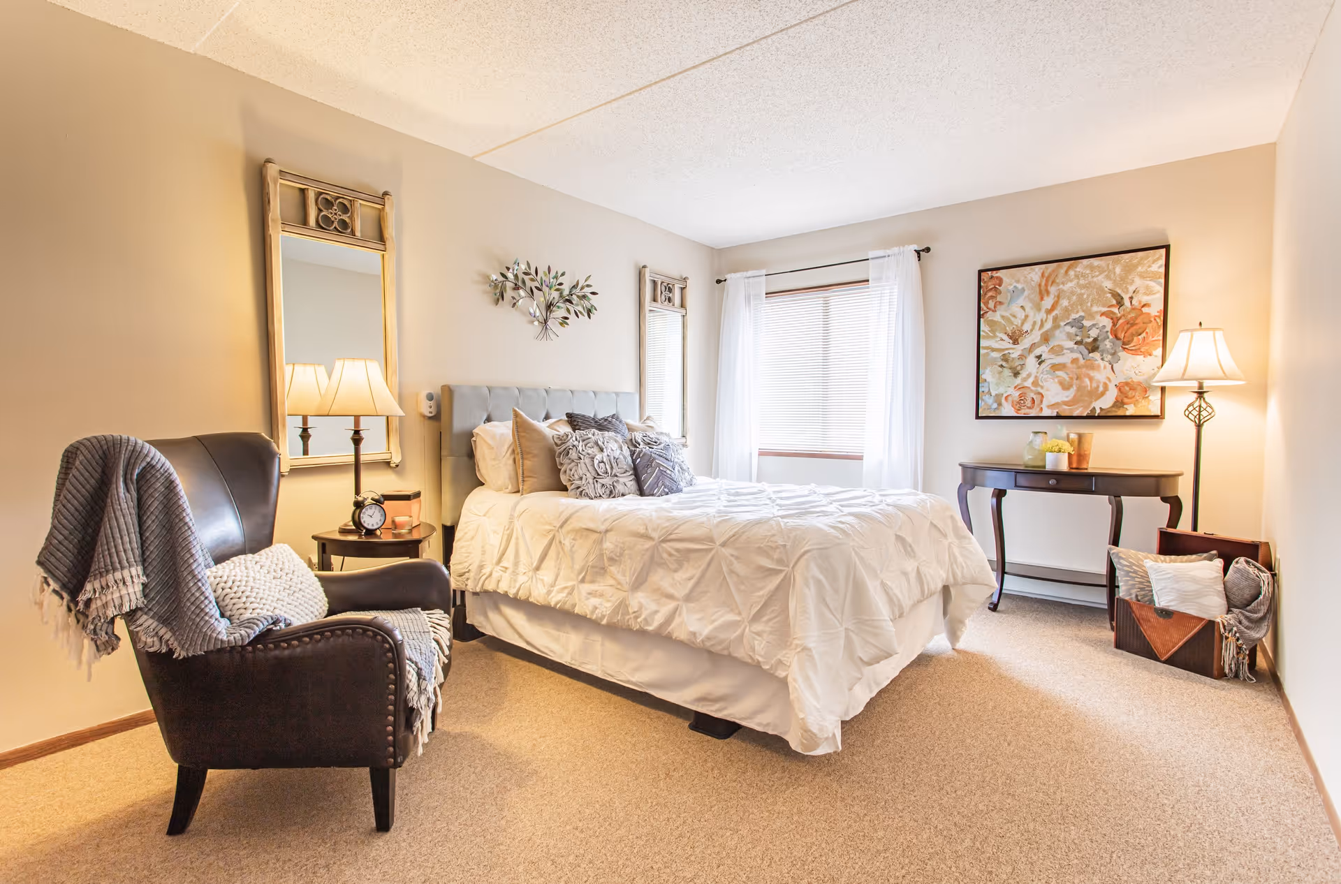 A cozy bedroom with a neatly made bed featuring white bedding and decorative pillows. To the left of the bed is a dark armchair draped with a gray blanket and a white pillow. A small round side table with a lamp and an alarm clock is next to the chair. On the wall above the bed are two decorative mirrors and a metal wall art piece. A window with white curtains allows natural light into the room. On the right side, there is a dark wooden console table with a lamp and decorative items, and a basket with pillows and blankets on the floor. The room has beige carpet and light-colored walls.