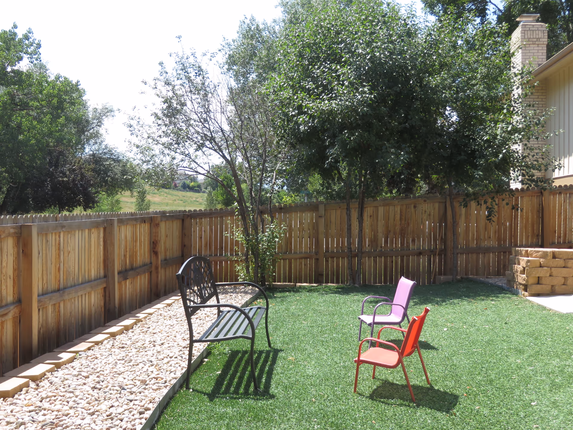 Fenced backyard with artificial grass, a metal bench, two colorful chairs, trees, and a gravel border.