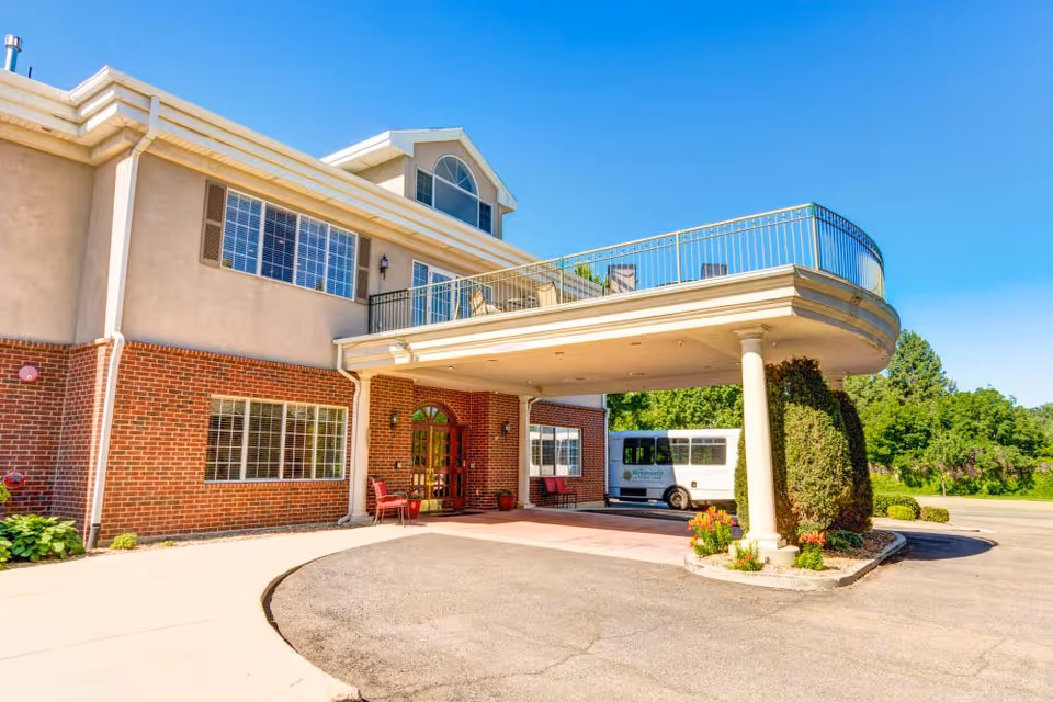 Exterior view of a senior living facility building with a covered entrance supported by columns. There are red chairs near the entrance door, a small garden with flowers and shrubs, and a white shuttle bus parked nearby. The sky is clear and blue.