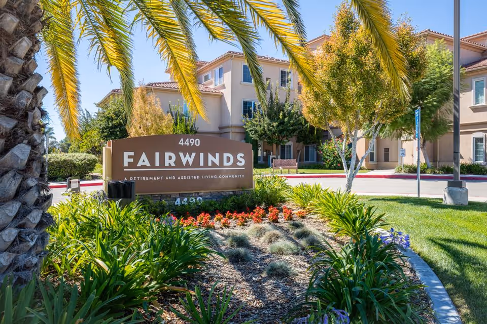 Entrance sign reading 'Fairwinds' surrounded by landscaped flower beds and palm trees with the facility building in the background.
