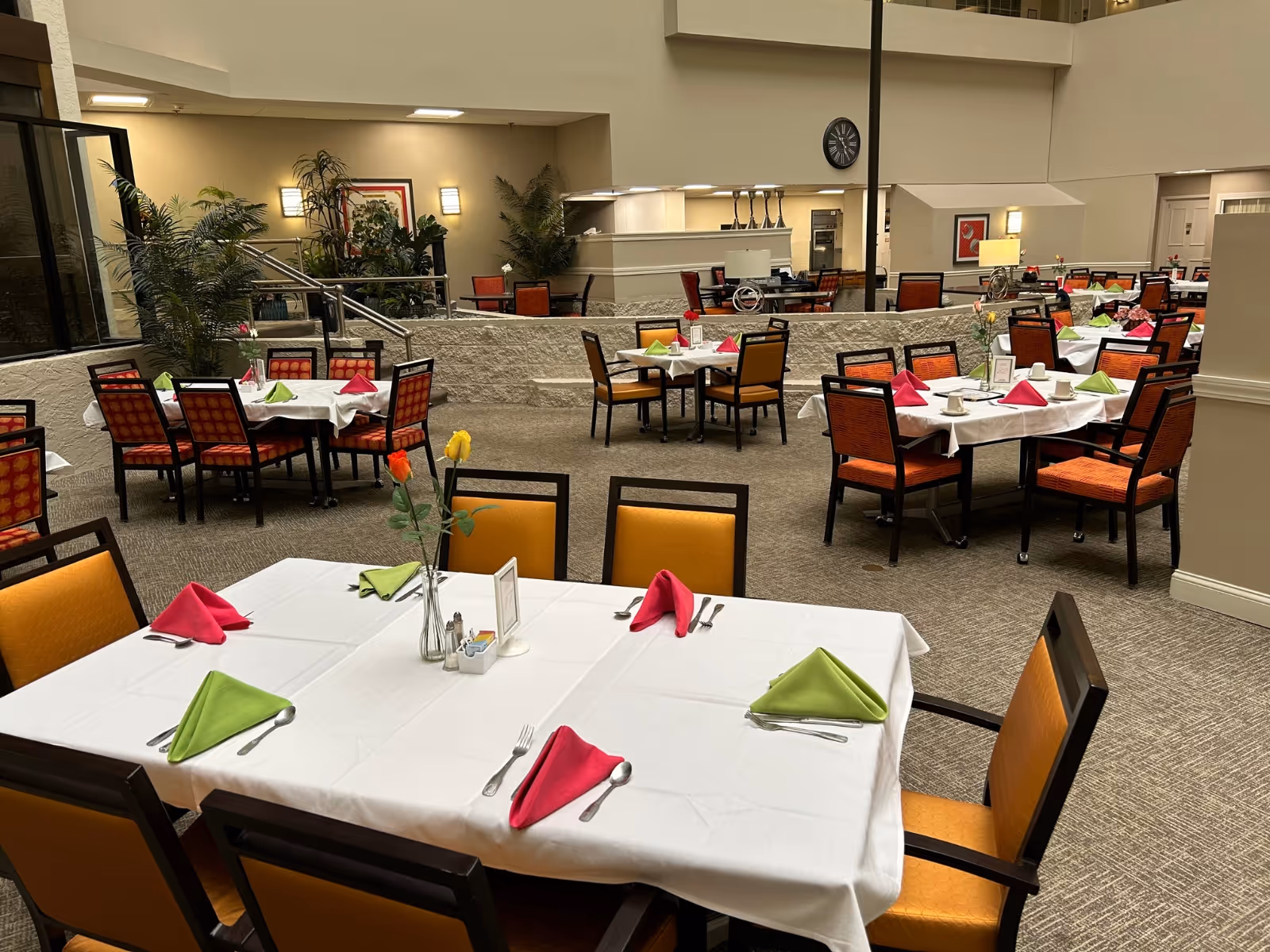 A spacious dining room in a senior living community with multiple tables covered in white tablecloths, each set with green and pink folded napkins, silverware, and small flower vases. The room has orange cushioned chairs, carpeted floors, and plants along the walls, with warm lighting and a clock on the wall.