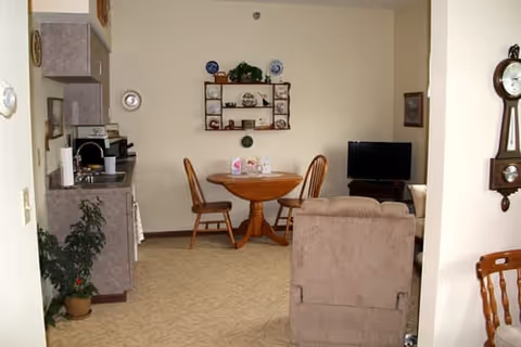 A cozy living area in a senior living facility featuring a small round wooden dining table with two chairs, a beige recliner chair, a flat-screen TV on a stand, and a wall-mounted shelf with decorative plates and plants. The room has light-colored walls and carpeted flooring, with a grandfather clock mounted on the right wall and a kitchenette visible on the left side.