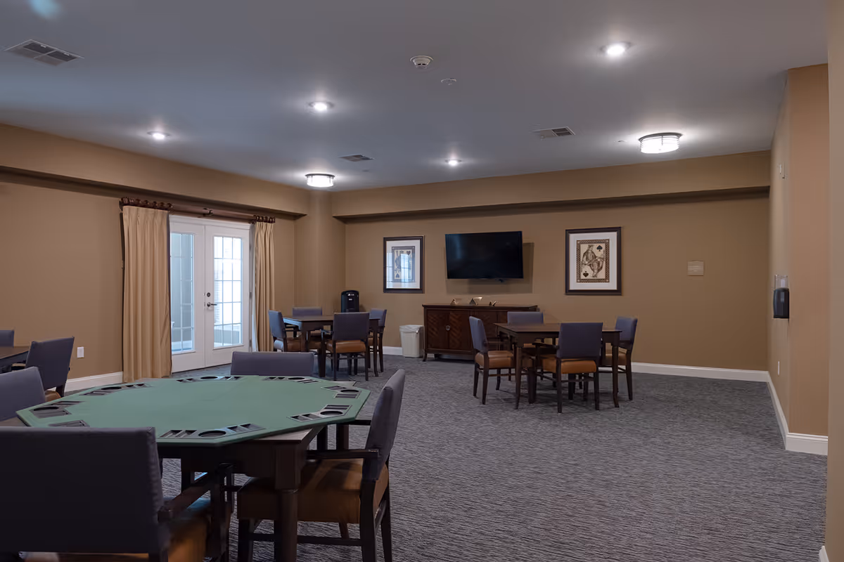 A spacious room with multiple tables and chairs arranged for group activities or dining. The room has beige walls, a carpeted floor, and ceiling lights. There is a wall-mounted TV between two framed pictures, and a set of French doors with beige curtains on one side.