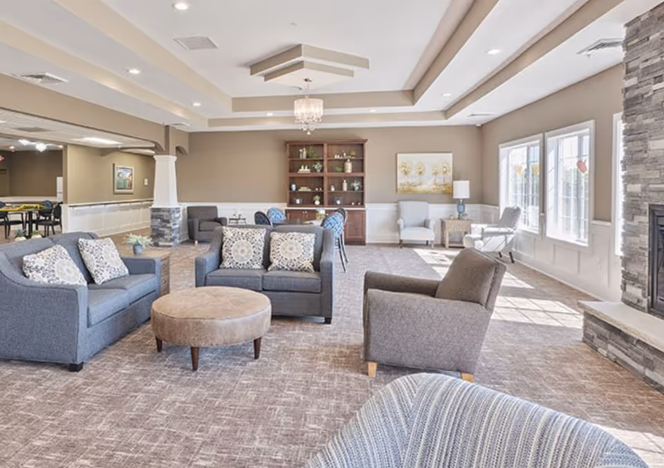 A bright and spacious assisted living facility common area with multiple seating options including gray sofas with patterned pillows, armchairs, and a round ottoman. The room features large windows allowing natural light to fill the space, a stone fireplace on the right, a wooden bookshelf against the far wall, and a ceiling with recessed lighting and a chandelier.