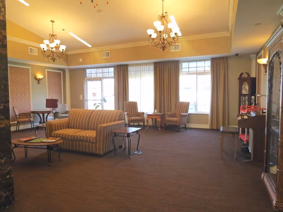 A spacious living room area with striped upholstered sofas and armchairs arranged around small wooden tables. The room features large windows with beige curtains, two chandeliers hanging from the ceiling, wall sconces, and a grandfather clock in the corner. The carpet is brown, and the walls are painted in warm neutral tones.