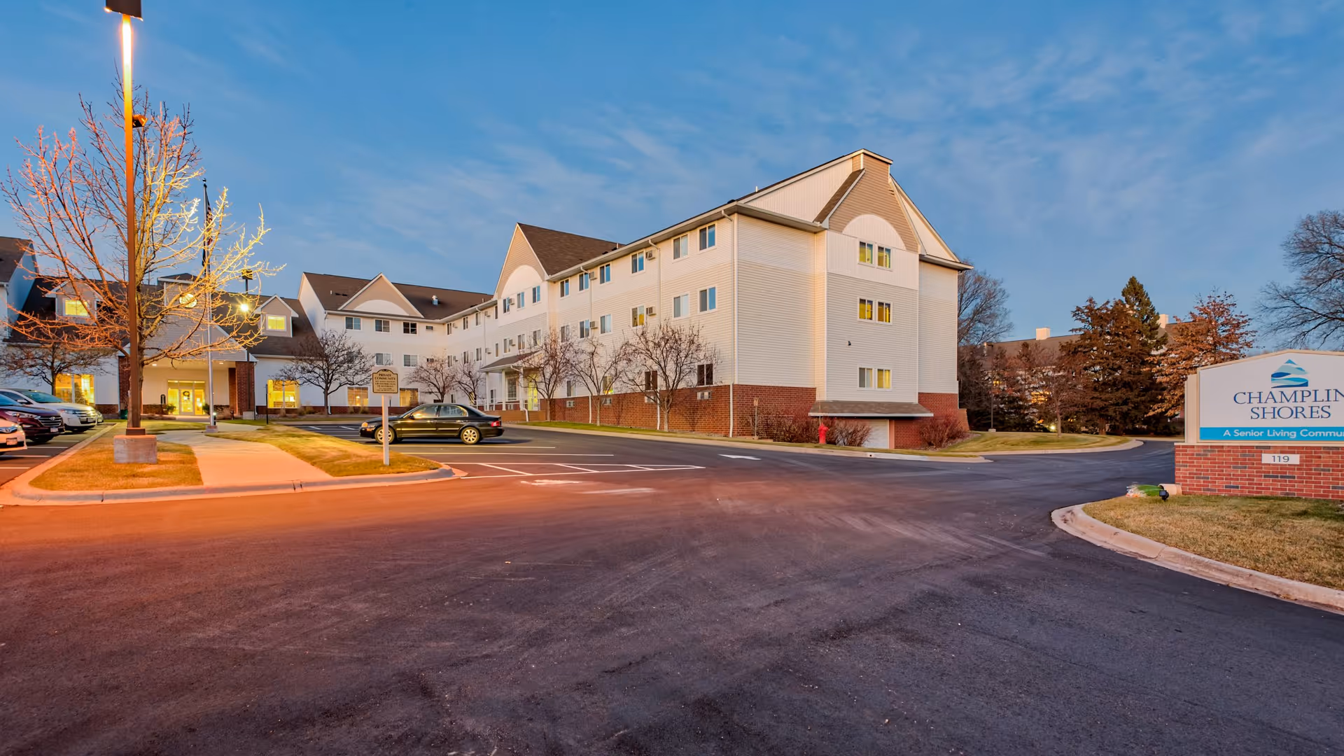 Exterior view of a multi-story senior living facility building with a parking lot in front, illuminated by street lamps at dusk. There are several cars parked, leafless trees, and a sign that reads 'Champlin Shores A Senior Living Community.'