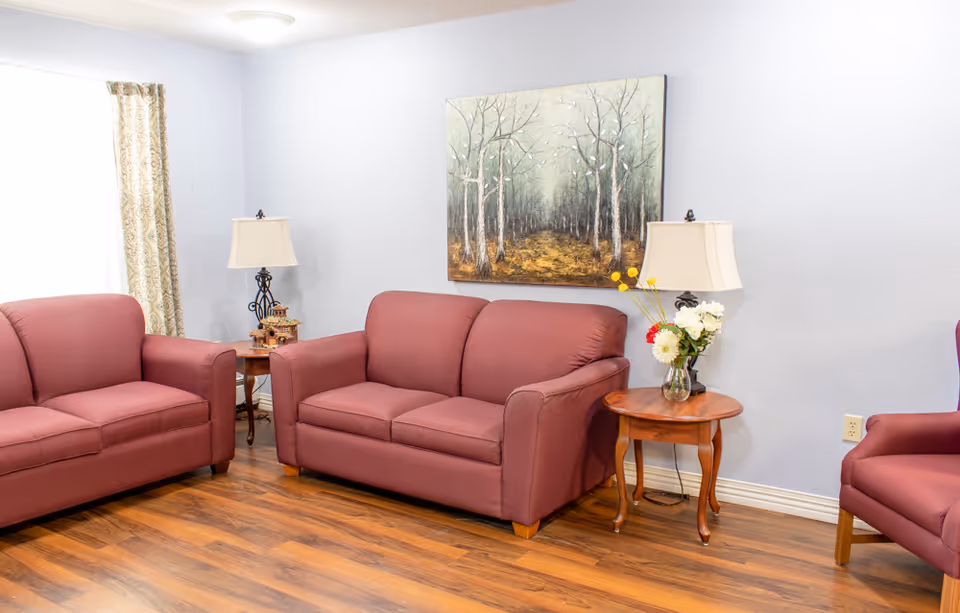 A cozy living room with two mauve sofas and a matching armchair arranged around two wooden side tables with lamps. A vase with flowers is on one table, and a painting of a forest scene hangs on the light blue wall. The room has wooden flooring and a window with patterned curtains.
