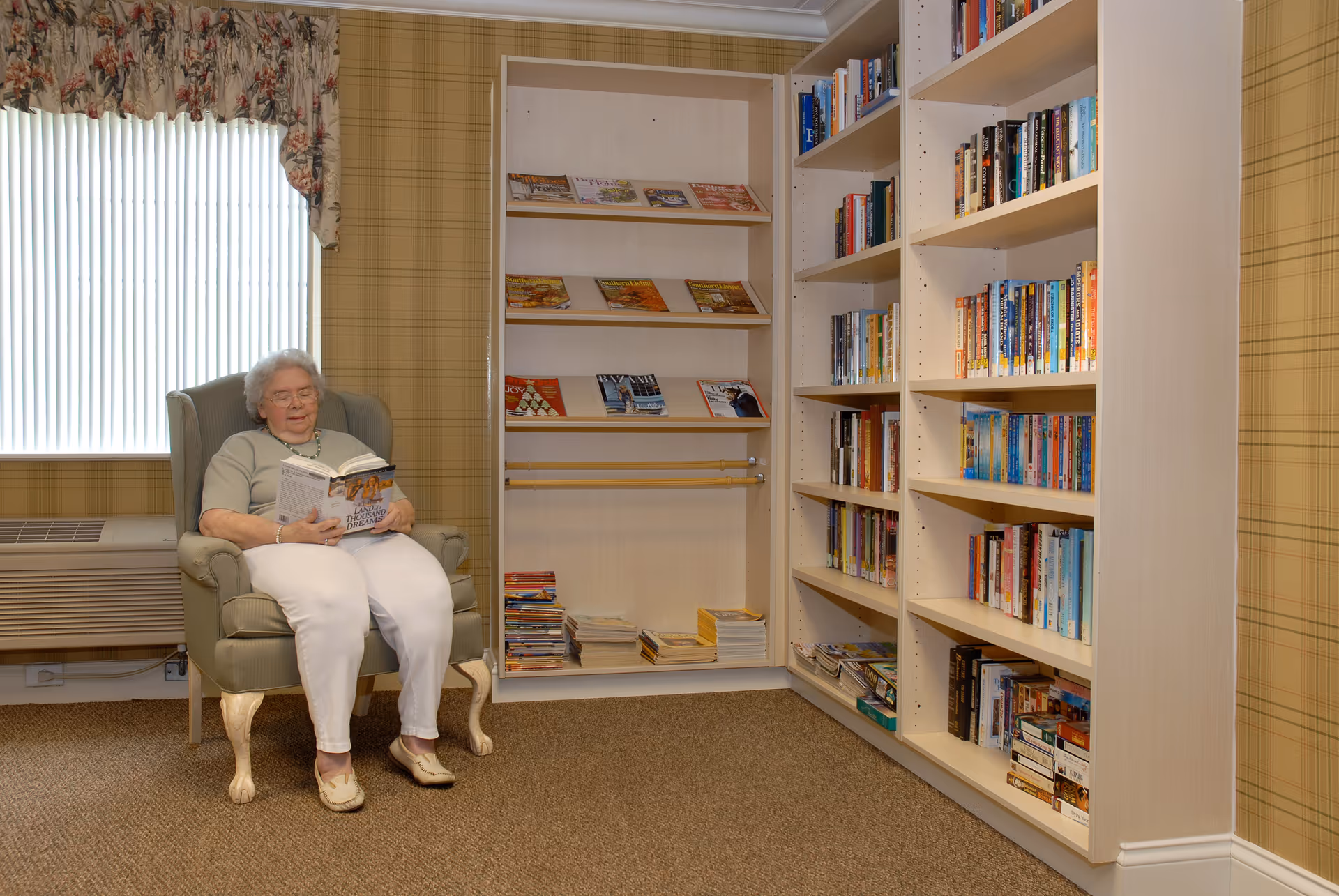 An elderly woman sitting in a cushioned armchair reading a book in a cozy room with bookshelves filled with books and magazines. The room has patterned wallpaper, a window with vertical blinds and floral valance, and carpeted floor.